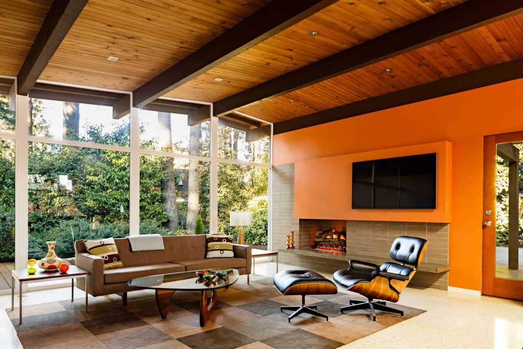 Spacious living room in a house with window wall, wooden ceiling, brown sofa and black armchair, brown brick fireplace and a TV above the fireplace on an orange wall